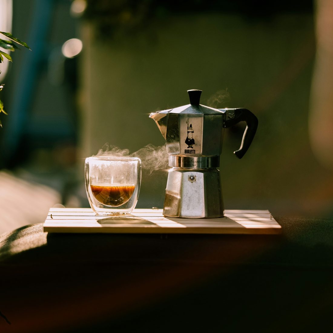 A Bialetti moka pot beside a steaming glass of fresh espresso