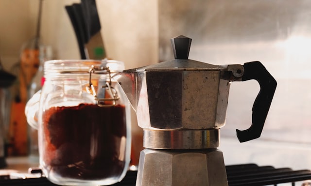 A moka pot on a hob beside a jar of ground coffee